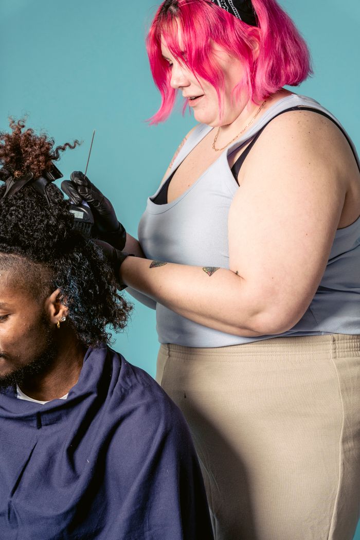Side view of professional hairdresser in black gloves coloring hair with brush to African American male client against blue background